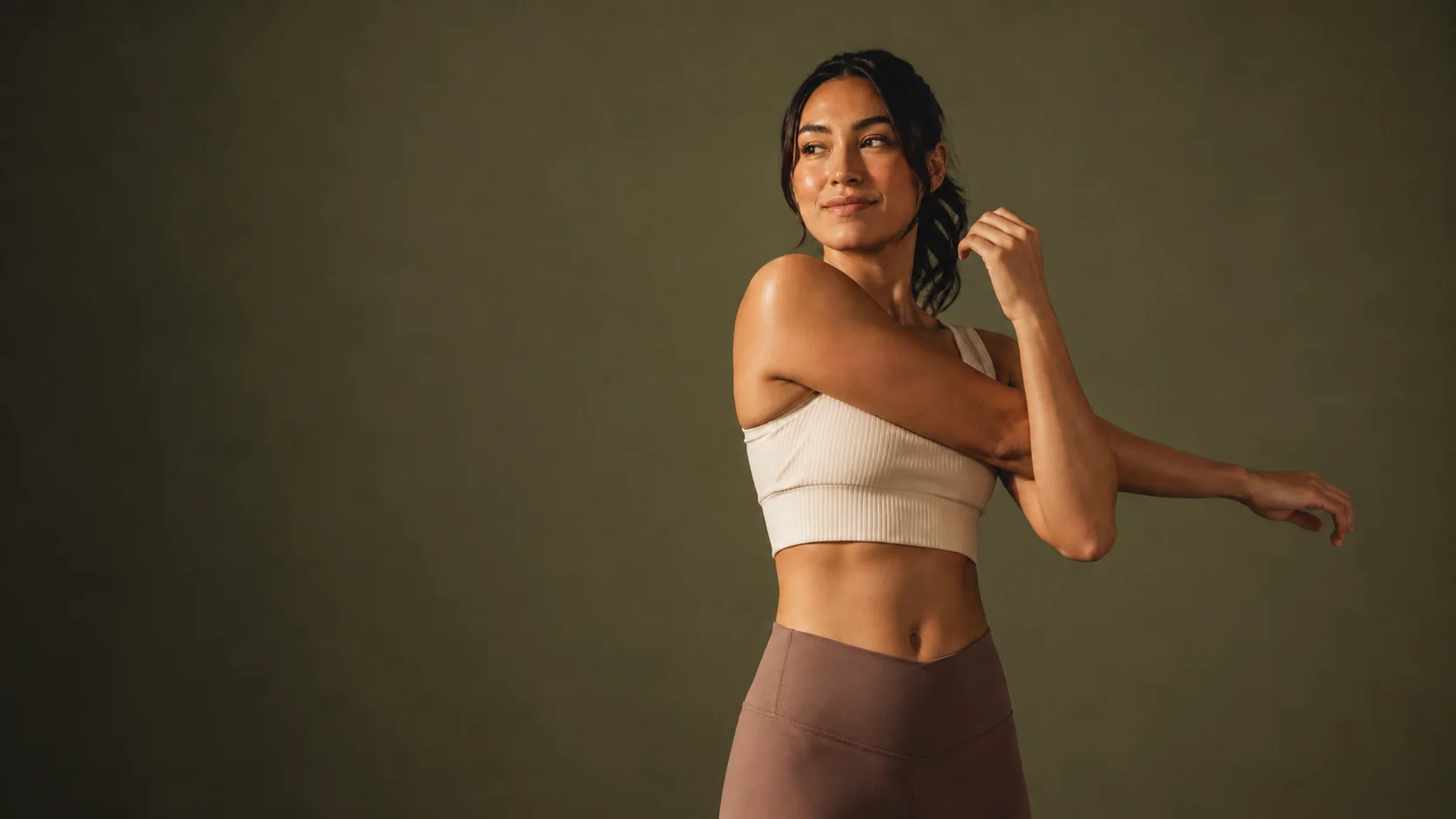 Mulher alongando os braços, em roupa de treino, fundo verde-oliva